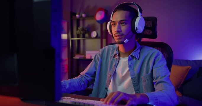 Happy asia guy gamer wear headphone participation play video game colorful neon lights computer in living room at night modern house. Esport streaming game online, Home quarantine activity concept.