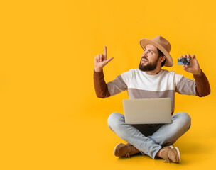 Handsome man with gift card and laptop pointing at something on yellow background. Black Friday sale