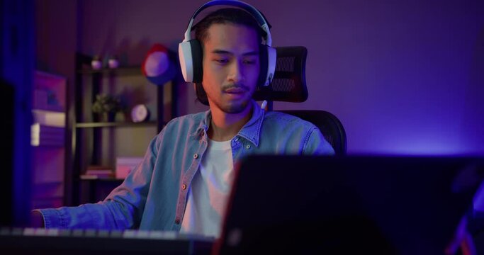 Happy asia guy gamer wear headphone participation play video game colorful neon lights computer and record a podcast on tablet in living room at night modern house. Esport streaming game online.