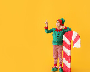 Cute little boy dressed as elf with candy cane pinata pointing at something on color background