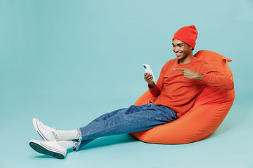 Full body young smiling cheerful fun happy african american man 20s wearing orange shirt hat sit in bag chair use hold mobile cell phone isolated on plain pastel light blue background studio portrait.
