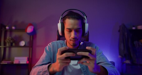 Happy asia man gamer wear headphone competition play video game online with smartphone colorful neon light in living room at night modern house. Esport streaming game online, Home quarantine activity. - Powered by Adobe
