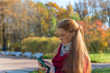 Fototapeta na wymiar Beautiful young woman with long red hair and freckles on her face with a mobile phone in a sunny autumn park