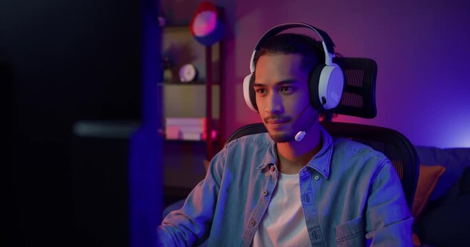 Unhappy asia guy gamer wear headphone participation play video game colorful neon lights computer in living room at night modern house. Esport streaming game online, Home quarantine activity concept.