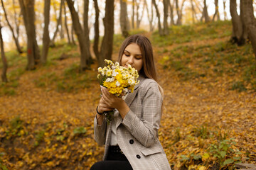 a young beautiful girl in a gray jacket is sitting on a bridge over a stream and holding a bouquet of autumn flowers in an autumn city forest park