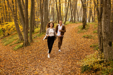 a young handsome guy in an autumn beige jacket runs through the autumn city forest park with a young beautiful girl in a gray jacket