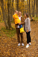 a young handsome guy in an autumn beige jacket is holding a little girl in a yellow raincoat next to a young beautiful girl in a gray coat autumn city park forest