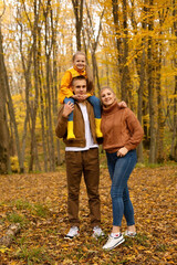Naklejka na ściany i meble a young handsome guy in an autumn beige jacket is holding a little girl in a yellow raincoat next to his young mother in a brown knitted sweater autumn city park forest