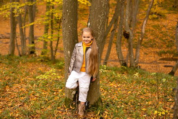 a beautiful blonde girl in a yellow sweater and a light jacket stands by a tree in the urban autumn forest