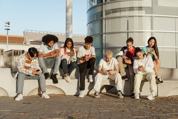 Multicultural group of young trendy friends using cellphones and social media - Students sitting in...