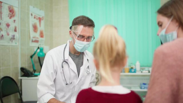 Zoom in rack focus shot of male doctor in protective face shield and mask talking to young mother visiting pediatrician with little daughter recovering from coronavirus disease