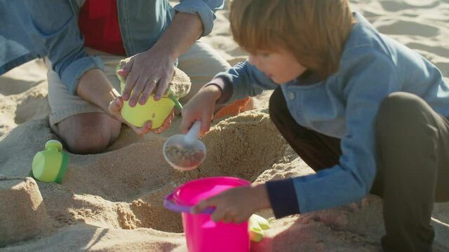 Kid picking sand in bucket with childrens shovel on beach. Cute Caucasian little boy spending time with father while playing in sand on sea shore on summer day. Recreation, holiday, family concept