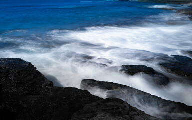 Smooth silky waves flow over the black rocks on the beach. Panoramic motion blur rocky shore image.