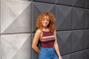 Fototapeta premium young redhead with curly hair and jeans smiling in the city with a gray wall in the background