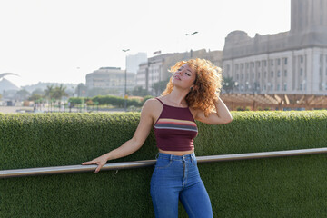 young redhead with curly hair and jeans shaking her hair leaning on a wall of green grass with sun behind and the city skyline.