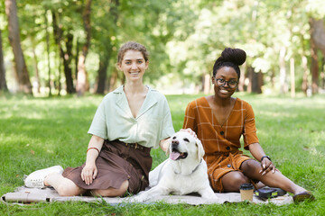 Full length portrait of two young women sitting on grass in park with dog outdoors and looking at camera, copy space © Seventyfour