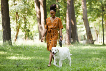 Full length portrait of young African-American woman walking dog in park outdoors in Summer, copy...