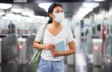 Asian woman in face mask standing in subway with metro ticket gates in background.