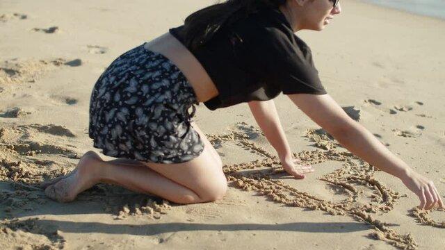 Side view of pensive teenage girl drawing heart in sand on beach. Long shot of cheerful girl wearing T-shirt and skirt sitting on sea shore on hot summer day. Recreation, seaside resort concept