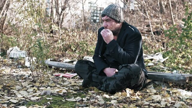 a hungry homeless unshaven man on the street in a torn suit smokes a cigarette an old beggar wants to eat a refugee without documents is sitting on the ground..