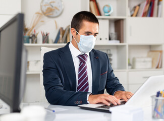 Professional business man in protective medical mask using laptop at workplace in office. High quality photo