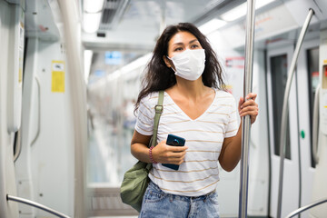 Asian woman in face mask with shoulder bag standing in subway car and using her smartphone.