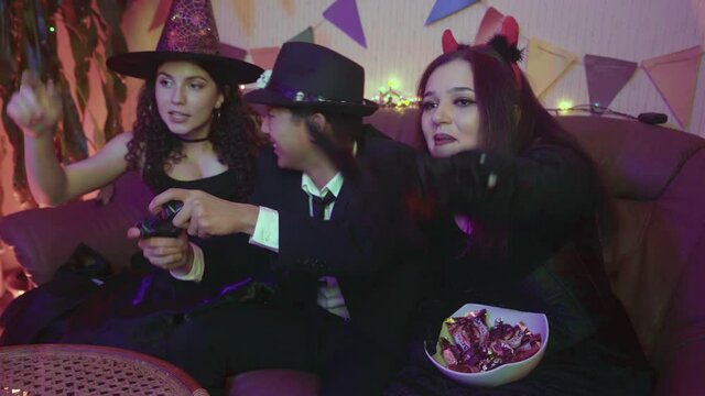 A guy in a creepy costume is actively playing on the console, and the girls happily support him at a Halloween party.