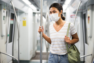 Black-haired asian woman in mask standing inside subway car and holding handrail.