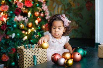 Fototapeta na wymiar a little African-American girl in a pink dress takes Christmas balls out of the box to decorate the Christmas tree.