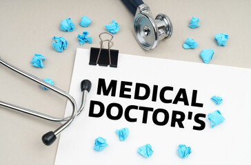 On the table is a stethoscope, a pen, blue crumpled pieces of paper and a sign with the inscription - MEDICAL DOCTORS - Powered by Adobe