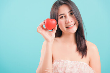 Obraz na płótnie Canvas Asian happy portrait beautiful young woman standing smile holding red heart on hand near face on blue background with copy space for text