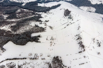 Aerial winter landscape with small rural houses between snow covered forest in cold mountains.