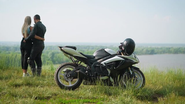Motorcyclist with slim girlfriend stands near contemporary bike on steep hill against picturesque lake on spring day backside view slow motion