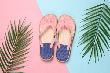 Colored flip flops with green palm leaves on blue-pink pastel background. Beach vacation concept....