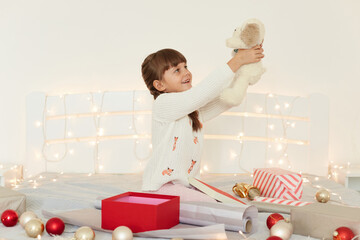 Side view portrait of charming kid wearing white sweater raised arms with soft toy dog while sitting on bed with Christmas decoration and garland, Happy new year.