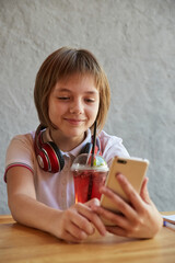 schoolgirl with headphones and red icy lemonade holds smartphone in hands. little girl using mobile phone at cafe. modern communication technology, using digital devices, gadgets, social networks