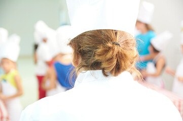 young cooking teacher in a white chef's hat with her back turned teaching a fun healthy cooking class to children