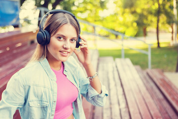 Image of a young stylish woman in youth fashionable clothes listening to music with headphones while sitting in the stands outdoors