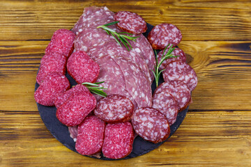 Different tasty sliced salami sausage on a black slate serving board on wooden table. Top view