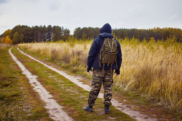 A man in a hooded jacket, camouflage trousers and a backpack on the background of an autumn road and forest.