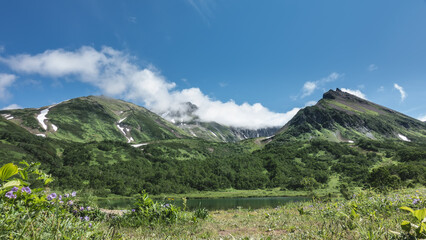A picturesque mountain range against the blue sky. There are patches of snow on the slopes. At the foot is the emerald lake. Green grass and wildflowers in the meadow. Kamchatka.