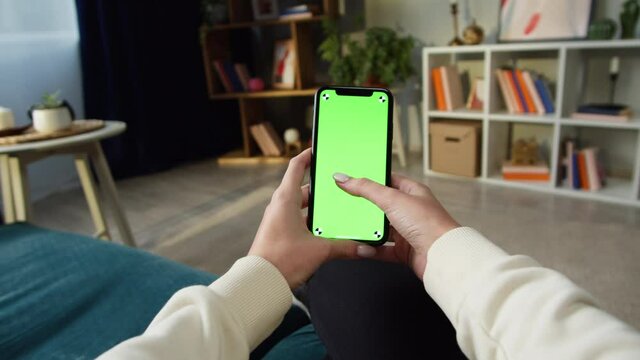 Woman using smartphone with green screen. Unrecognizable female person swiping in mobile phone with chroma key, sitting on sofa in living room. Online shopping, surfing internet. Spare time at home.