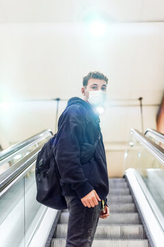 Young man with a backpack and white mask climbing an escalator in a metro station and looking at the camera