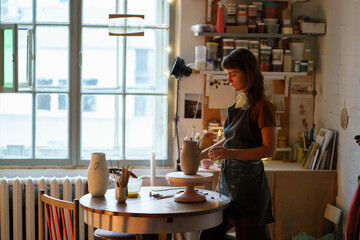 Ceramist at work: young self employed woman in apron creating potter jug for studio or craft kitchenware shop. Pottery master working with raw clay in workshop. Art hobby or small business concept