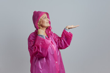 A mature smiling blonde in a magenta raincoat posing in a hood, extending her hand to the raindrops