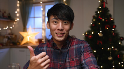 Close up portrait of Asian korean happy man looking camera speaking on video call with friend and having good talk sending greetings sitting near Christmas tree in room. New Year concept