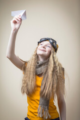 Youth Travel Concepts. Portrait of Positive Caucasian Teenage Girl in Flying Gear And Warm Hat Launching Origami Paper Plane Over Beige Background.