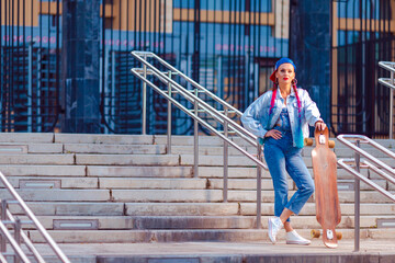Lovely Dreaming Caucasian Female Skateboarder Posing With Longboard At Fence on Stairways In Outdoor Environment While Holding Skateboard