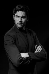 Fototapeta premium Black And White Portrait of Handsome Caucasian Brunet Businessman Wearing Black Suit Posing With Folded Hands Against Black Studio Background.