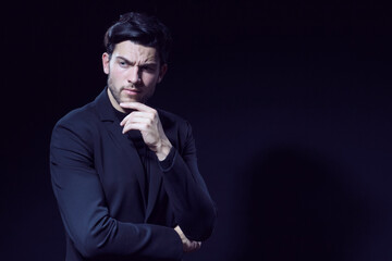 Fototapeta na wymiar Portrait of Handsome Caucasian Brunet Businessman Wearing Black Suit Posing With Folded Hands And Touching Chin Against Black Studio Background.